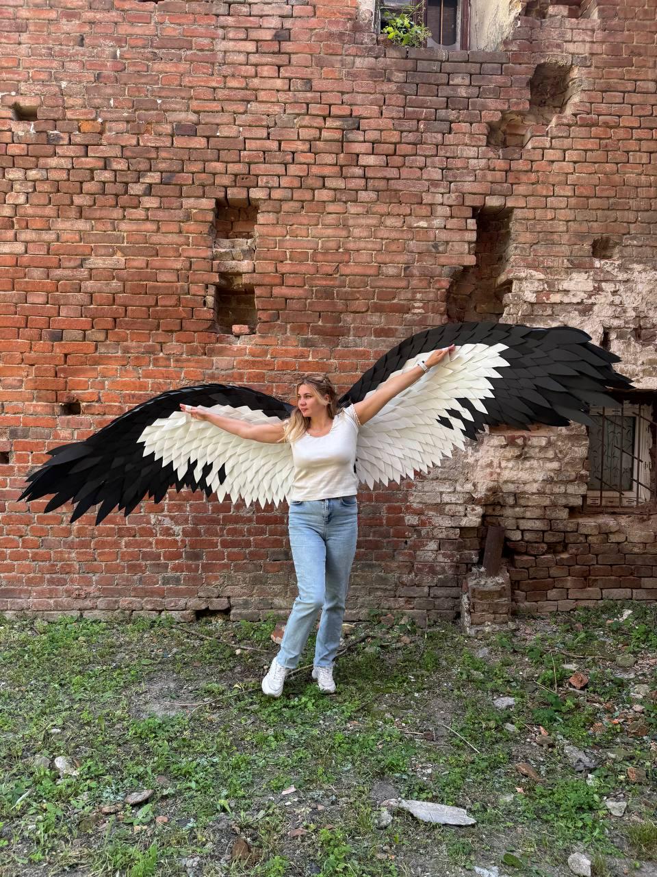 May include: A pair of large, wearable wings featuring a layered design with white inner feathers and black outer feathers. The wings are attached to a white short-sleeved top and light blue denim jeans, creating a wide, expansive span.