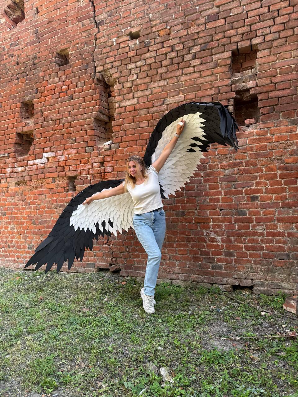 May include: Large, wearable bird-like wings featuring layered, pointed feathers in a high-contrast black and white color scheme. The wings are designed to be worn on the back, extending outward with a wide span, showcasing detailed feather textures.