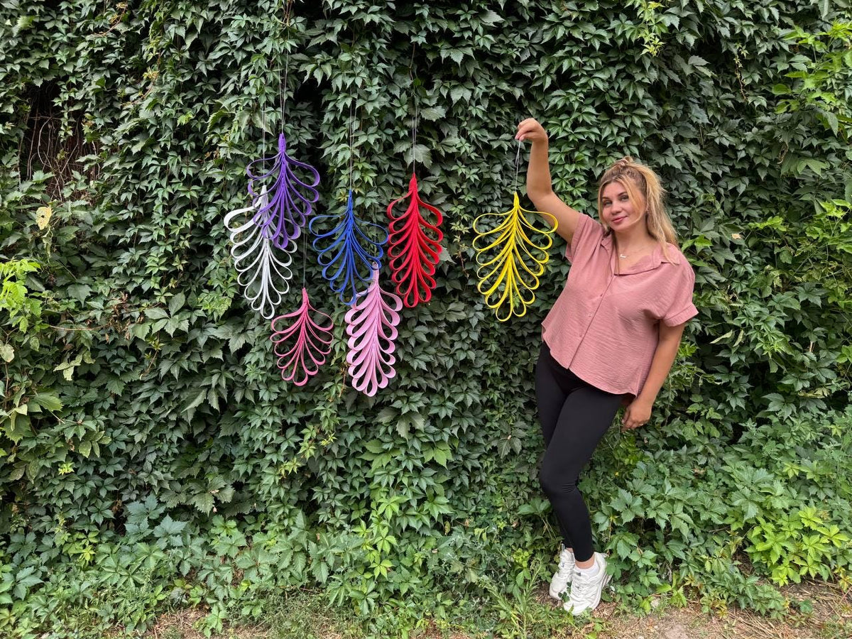 May include: A collection of six hanging leaf-shaped ornaments made from looped strips of material in purple, white, blue, red, pink, and yellow. Each ornament features a symmetrical, elongated design suspended by a thin string for display.