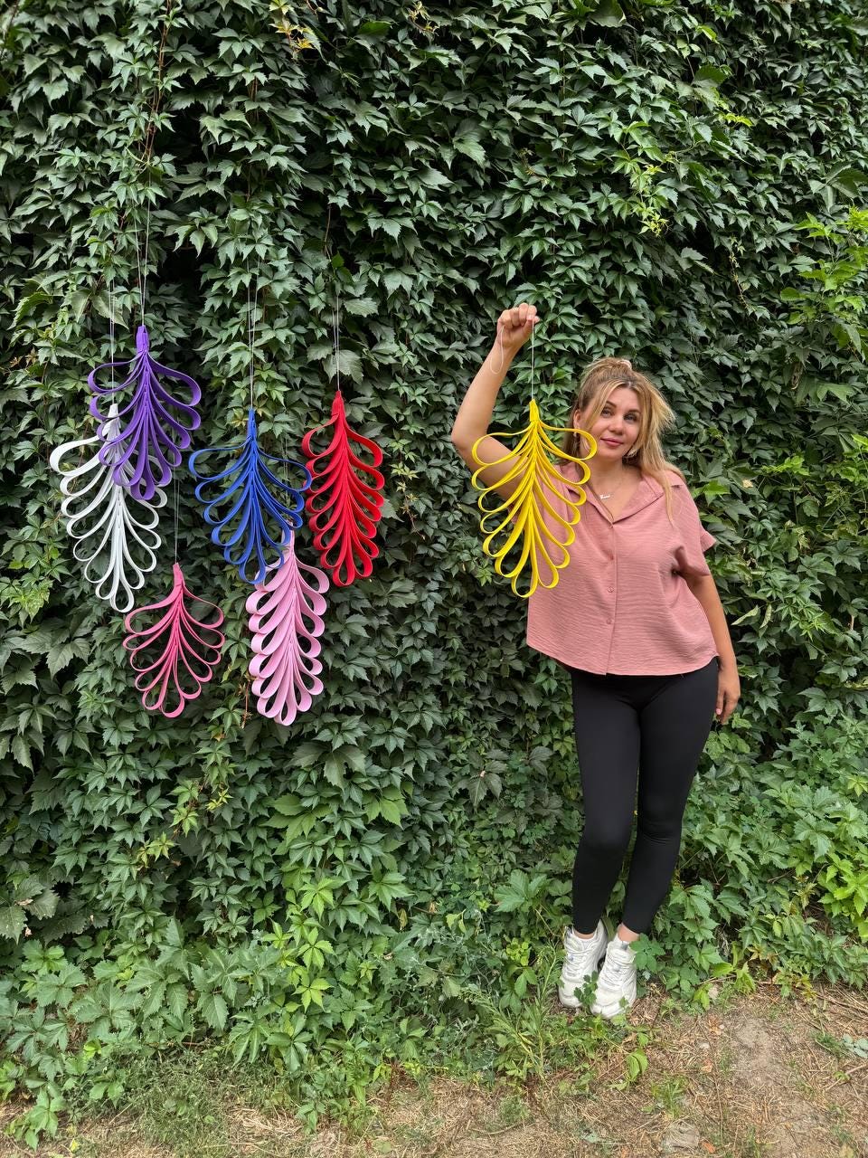 May include: A collection of hanging ornaments shaped like stylized leaves, crafted from looped strips of material in vibrant colors including purple, white, blue, red, pink, and yellow, suspended by thin strings against a wall of green foliage.