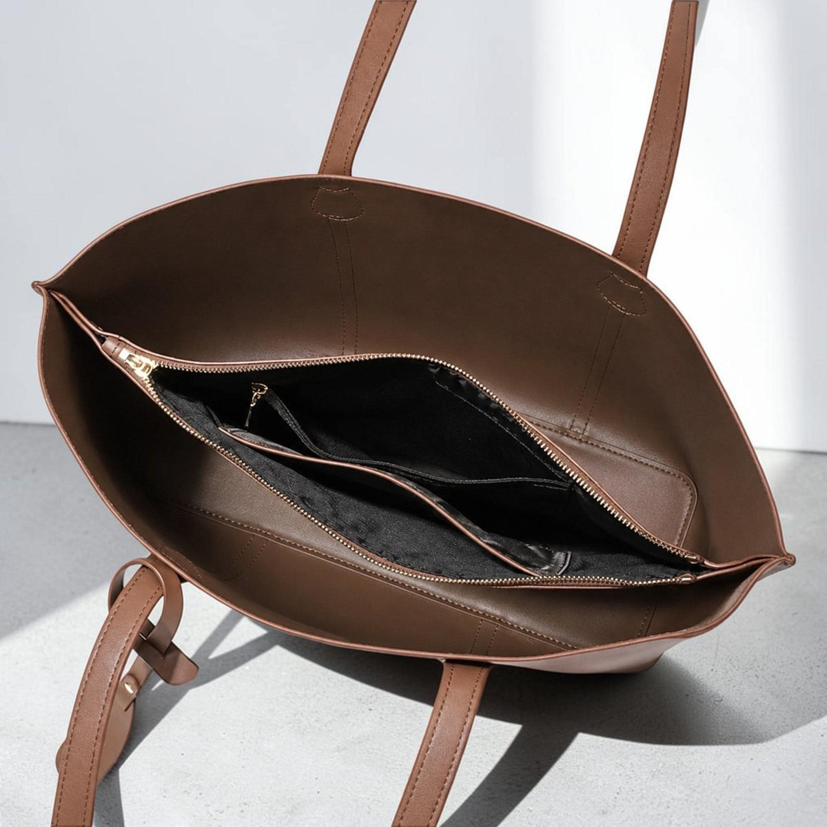 May include: A brown tote bag with two shoulder straps and an open top. The interior features a central zippered compartment lined in black fabric, which divides the main space into two sections, providing organized storage for personal items.