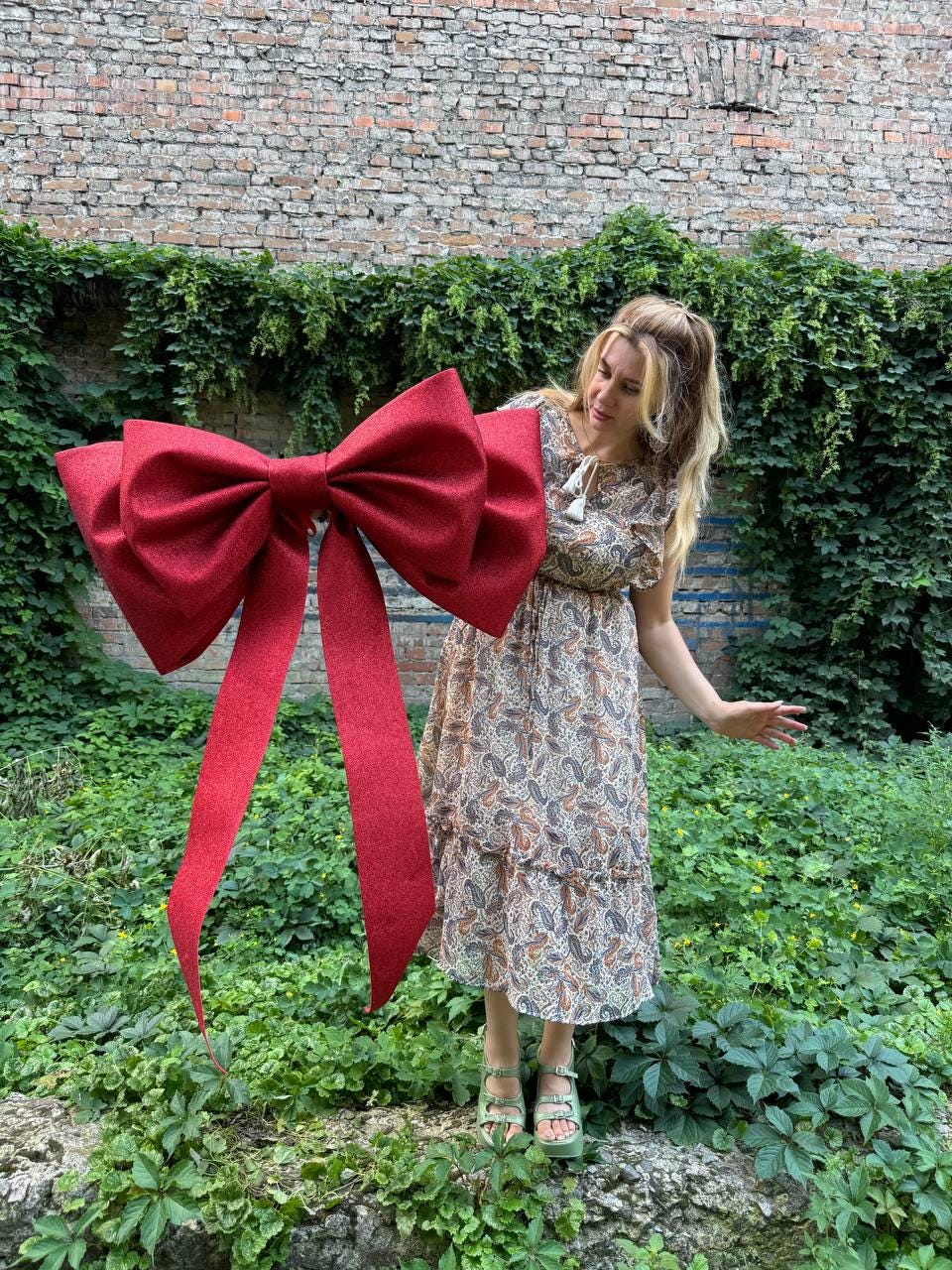 May include: A large, oversized decorative bow in a deep red, glittery fabric texture. The bow features two prominent loops and two long, trailing ribbon tails that extend downward. The material has a subtle, sparkling finish throughout.