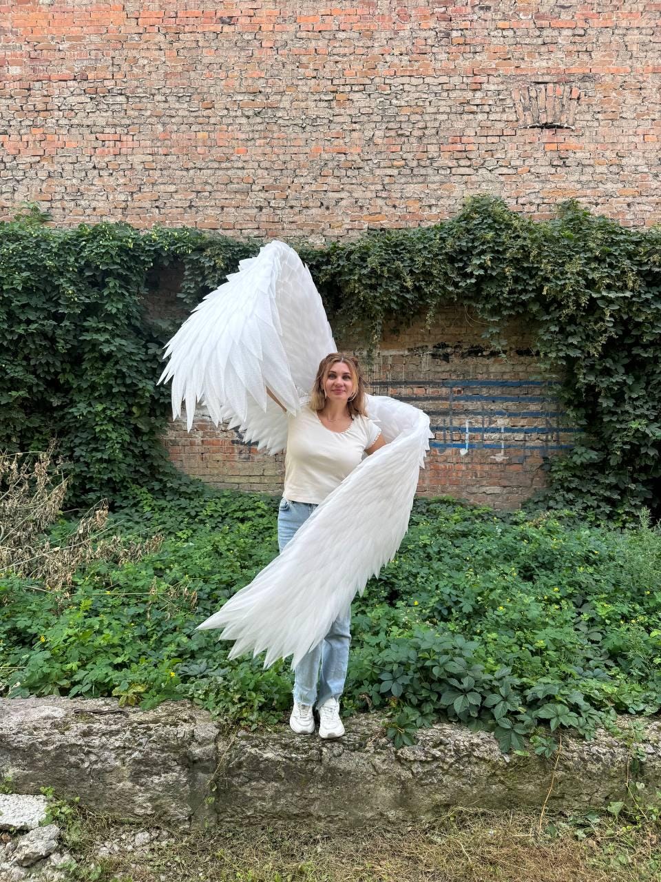 May include: Large white feathered wings attached to a harness, worn over a plain white short-sleeved t-shirt and light blue denim jeans. The wings feature layered, pointed white feathers that extend outward, creating a wide, expansive span.