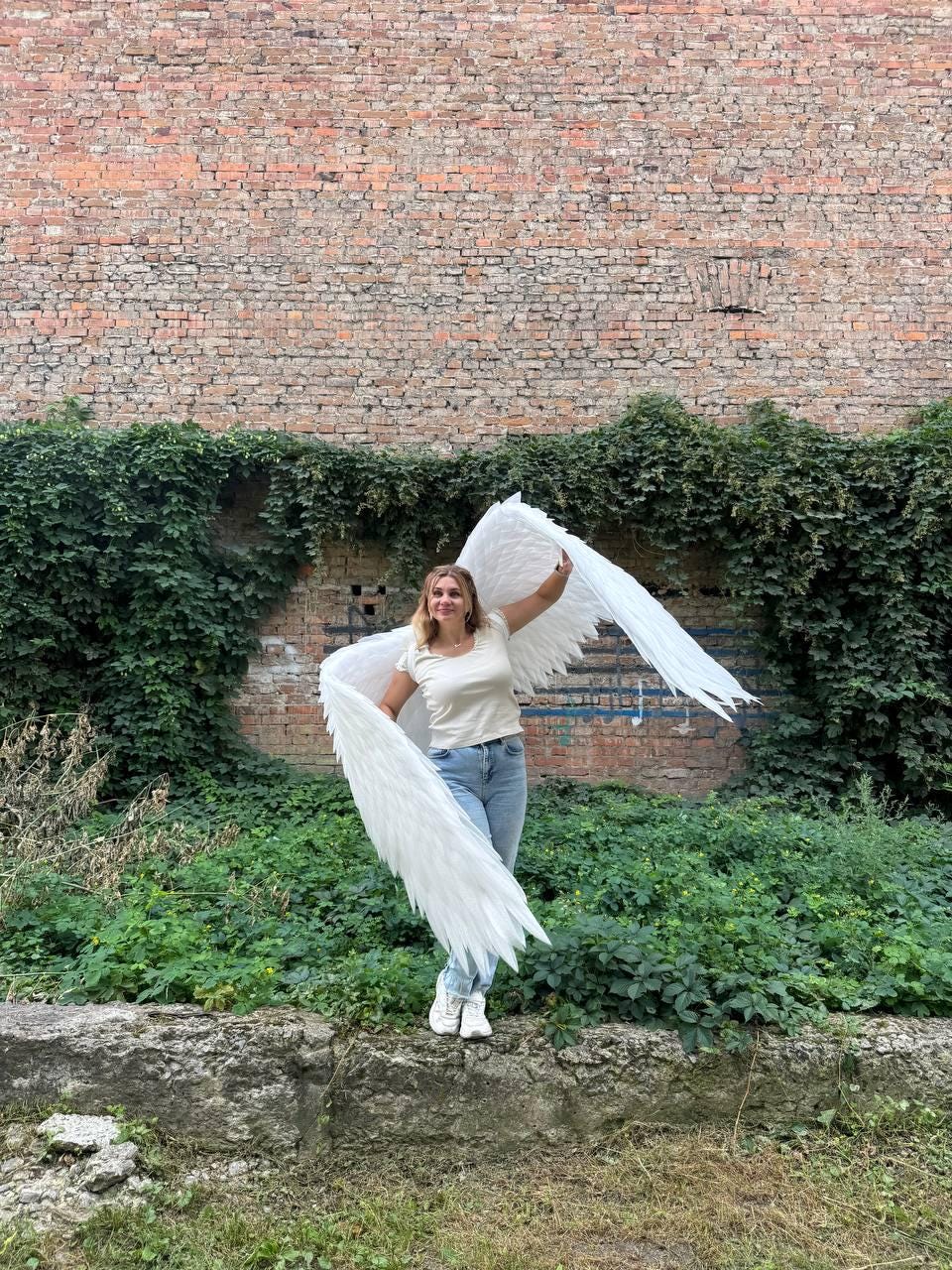 May include: Large, white, feathered angel wings attached to a harness, worn over a plain beige short-sleeved t-shirt and light blue denim jeans. The wings feature layered, pointed white feathers that extend outward, creating a wide, expansive span.
