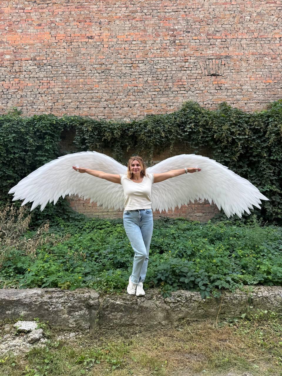 May include: Large, white, feathered angel wings attached to a white t-shirt and light blue denim jeans. The wings feature layered, pointed white feathers that extend horizontally, creating a wide span across the back of the garment.
