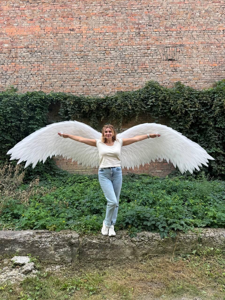 May include: Large, white, feathered angel wings attached to a white t-shirt and light blue denim jeans. The wings feature layered, pointed white feathers that extend horizontally, creating a wide span across the back of the garment.