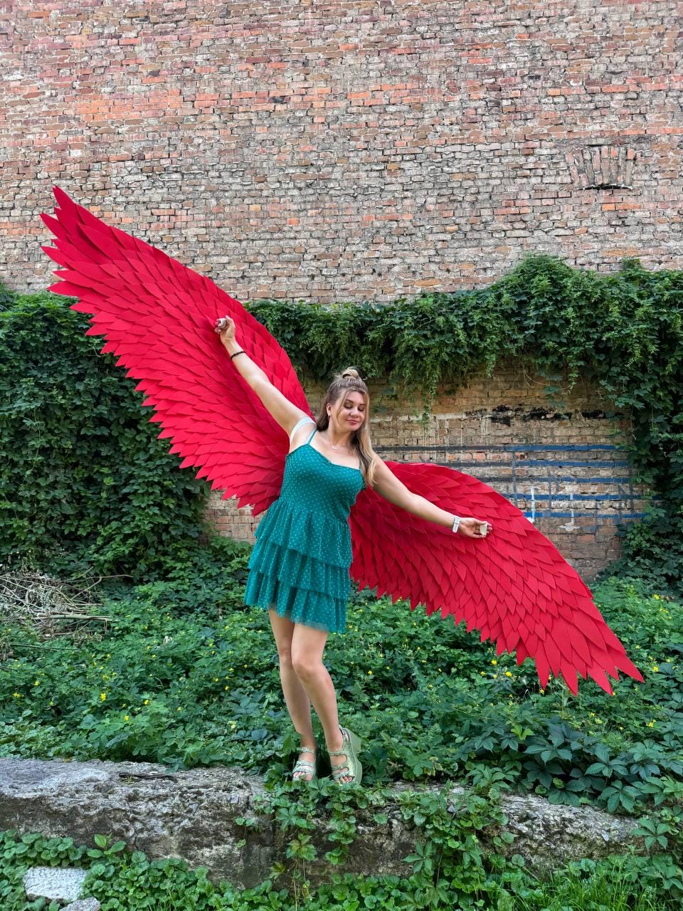May include: A pair of large, vibrant red feathered wings attached to a teal green sleeveless dress with a tiered skirt. The wings feature layered, pointed feather-like segments that extend outward, creating a wide, expansive span across the frame.