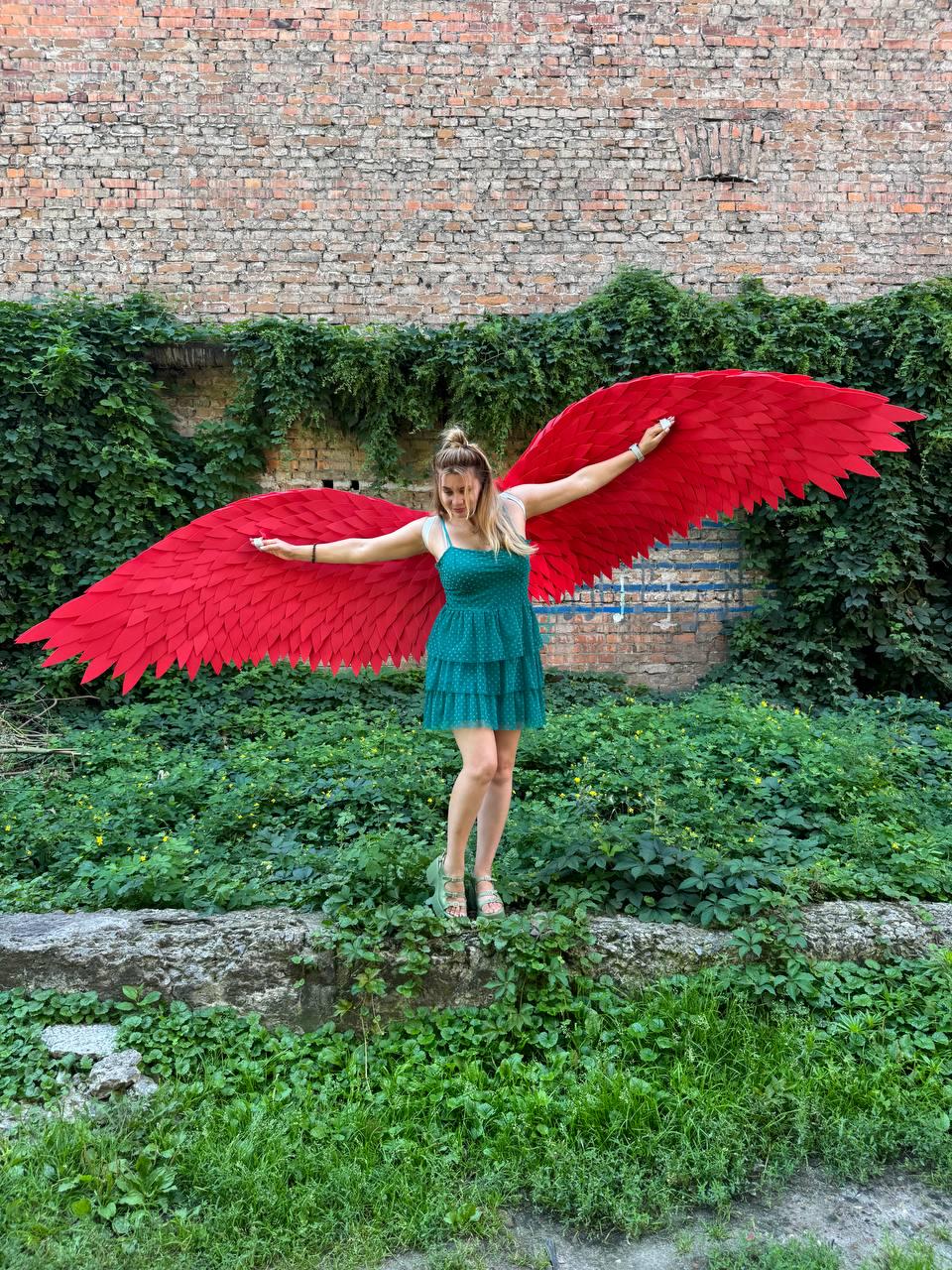 May include: A pair of large, vibrant red feathered wings attached to a teal, tiered, sleeveless mini dress with a subtle polka dot pattern. The wings feature layered, pointed feather-like segments that extend horizontally from the shoulders.