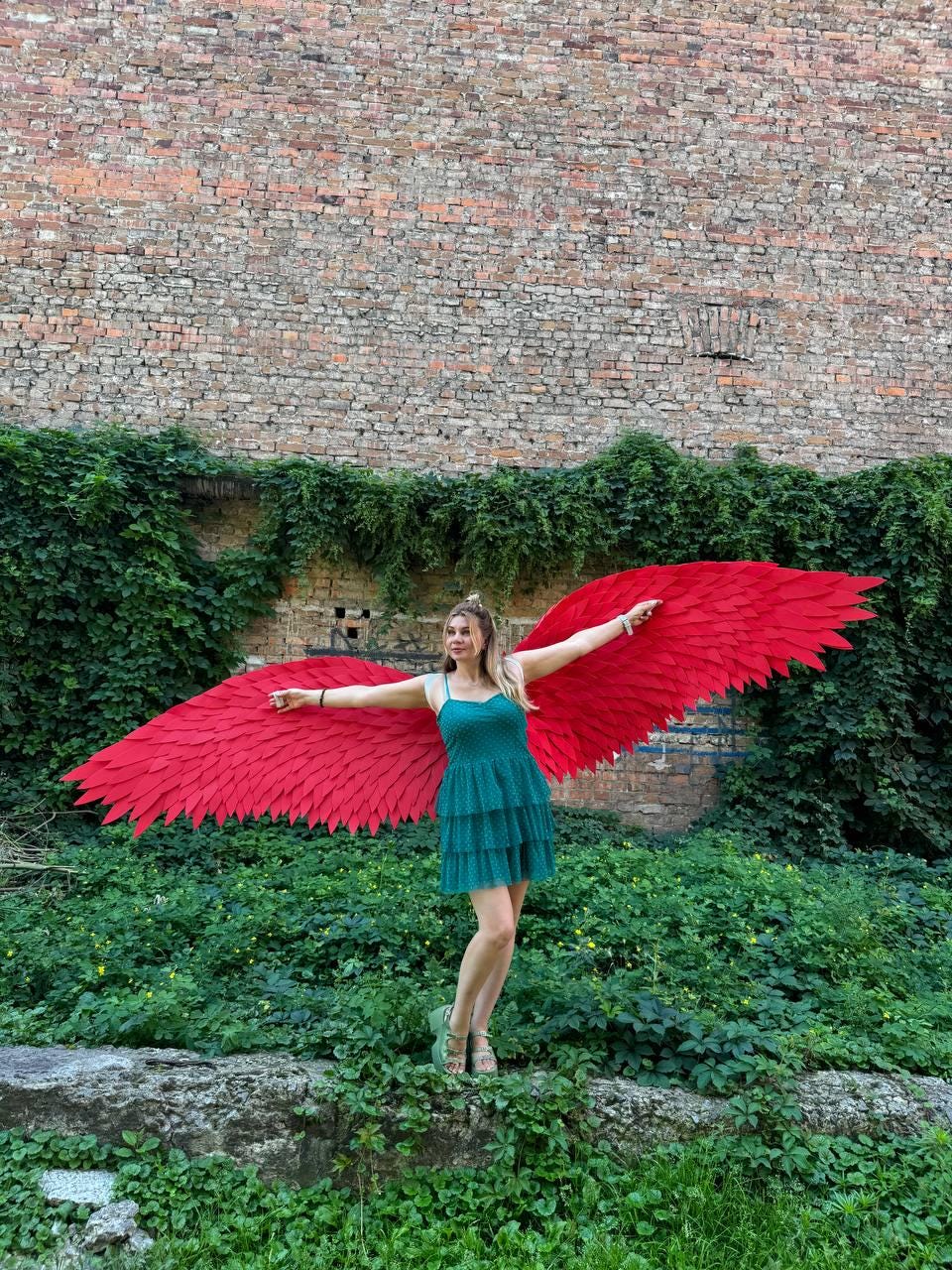 May include: A pair of large, vibrant red feathered wings attached to a teal, sleeveless, tiered mini dress. The wings feature a detailed, layered feather design that spans wide, while the dress has a fitted bodice and a textured, layered skirt.