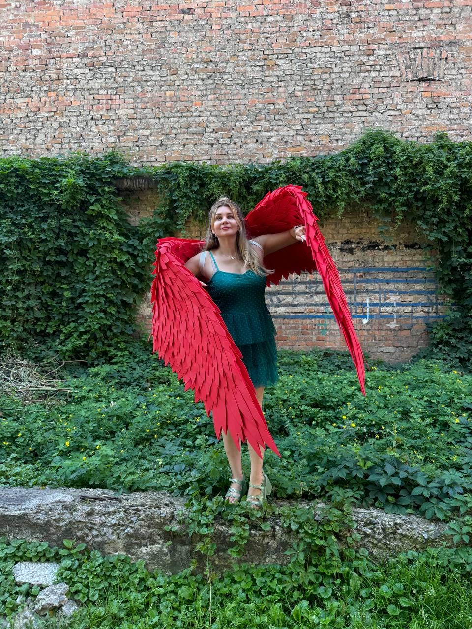 May include: A pair of large, vibrant red feathered wings with a layered, pointed design, worn over a dark green, sleeveless, tiered mini dress with thin shoulder straps and a subtle polka dot texture throughout the fabric.