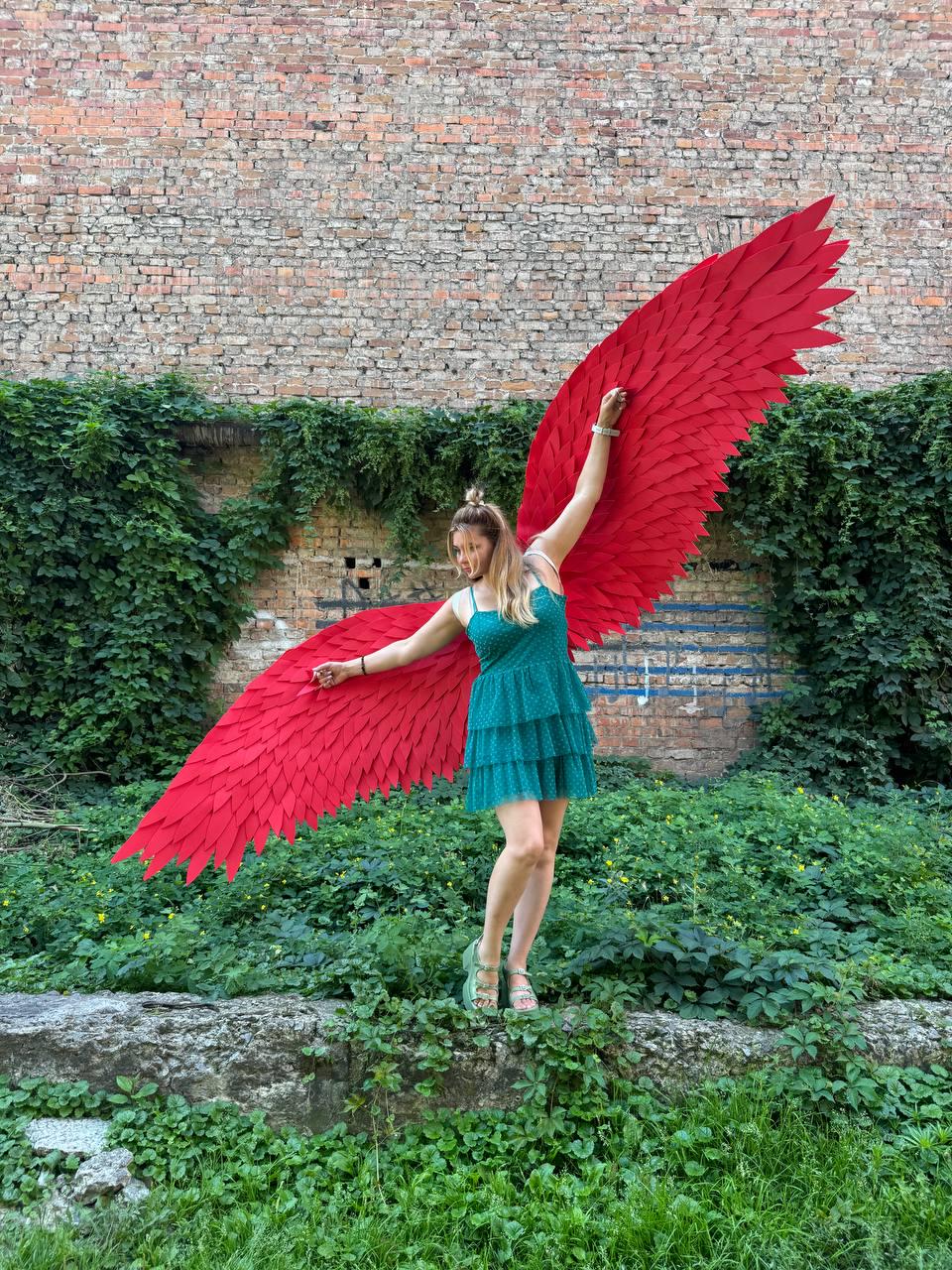 May include: A pair of large, vibrant red feathered wings attached to a teal, tiered, sleeveless dress. The wings feature a layered, pointed feather design that extends outward, while the dress has a textured, dotted fabric and a knee-length hem.