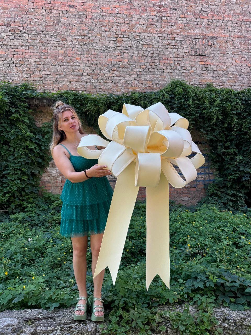 May include: A large, oversized decorative bow in a pale cream color. The bow features multiple voluminous loops arranged in a circular, multi-layered structure, with two long, wide ribbon tails hanging down from the center, ending in V-shaped notches.