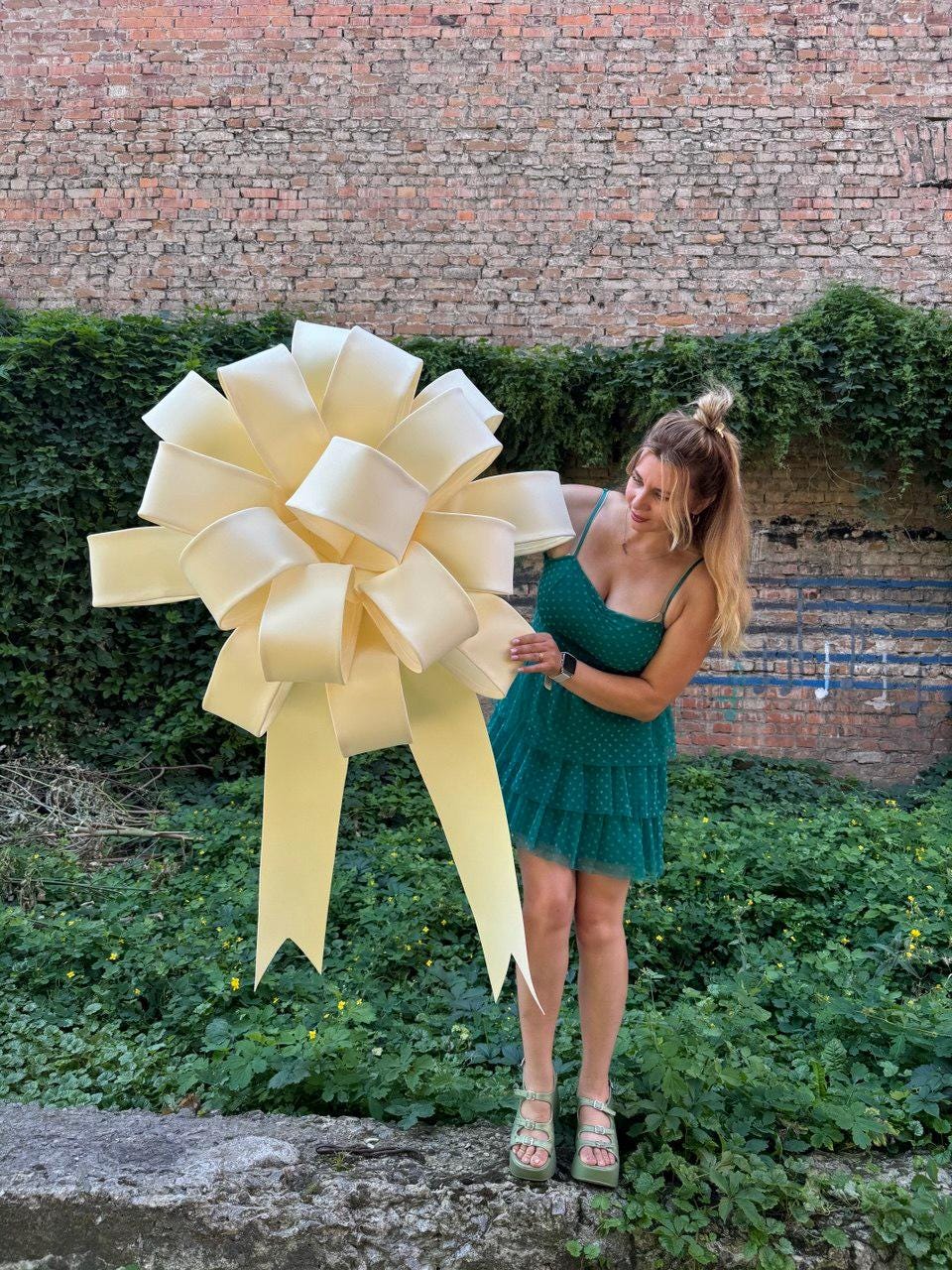 May include: A large, pale yellow decorative bow with multiple loops and two long, pointed tails. The bow is constructed from a smooth, solid-colored material and features a voluminous, rounded shape with symmetrical layers radiating from the center.