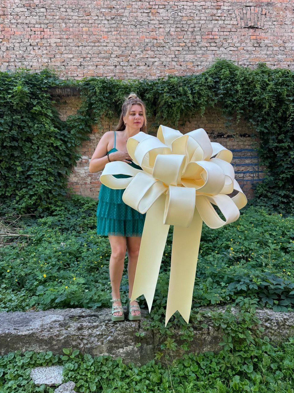 May include: A large, pale yellow decorative bow featuring multiple voluminous loops and two long, wide ribbon tails hanging downward. The bow is constructed from a smooth, matte material and is designed to be held or displayed as a statement piece.