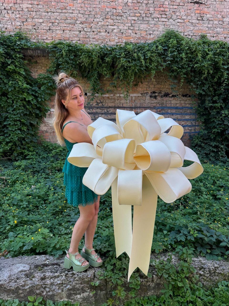 May include: A large, oversized decorative bow in a pale cream color, constructed from wide, smooth ribbon loops. The bow features a voluminous, multi-layered design with two long, trailing ribbon tails extending downward from the center knot.