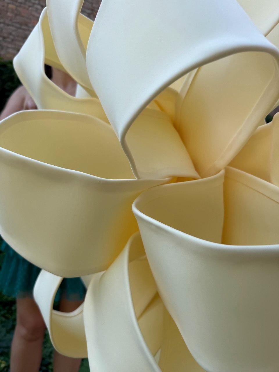 May include: A large, pale yellow decorative object constructed from multiple curved, interlocking loops of smooth material. The structure features a series of open, rounded segments that overlap to create a complex, three-dimensional floral-like shape.