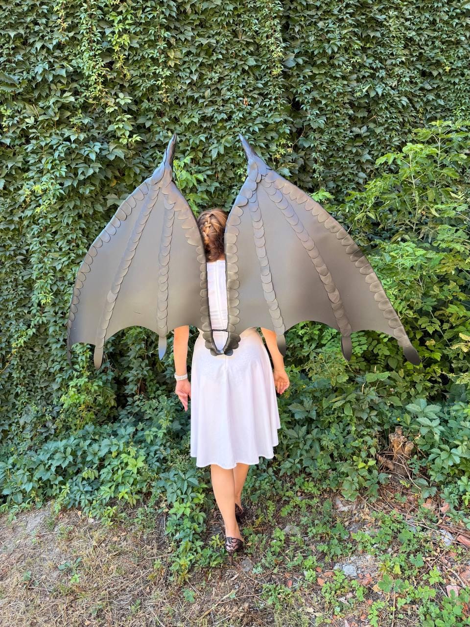 May include: A pair of large, dark gray dragon wings with a segmented, scale-like texture and pointed tips. The wings are attached to a harness worn over a plain white sleeveless dress. The structure features distinct ribbing and a wide, expansive span.