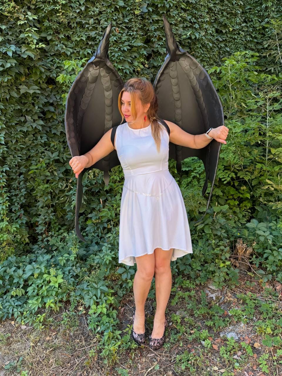 May include: A white sleeveless dress with a high-low hemline paired with large, black, textured dragon-style wings. The wings feature a segmented, scale-like pattern and are worn with black straps over the shoulders of the white dress.