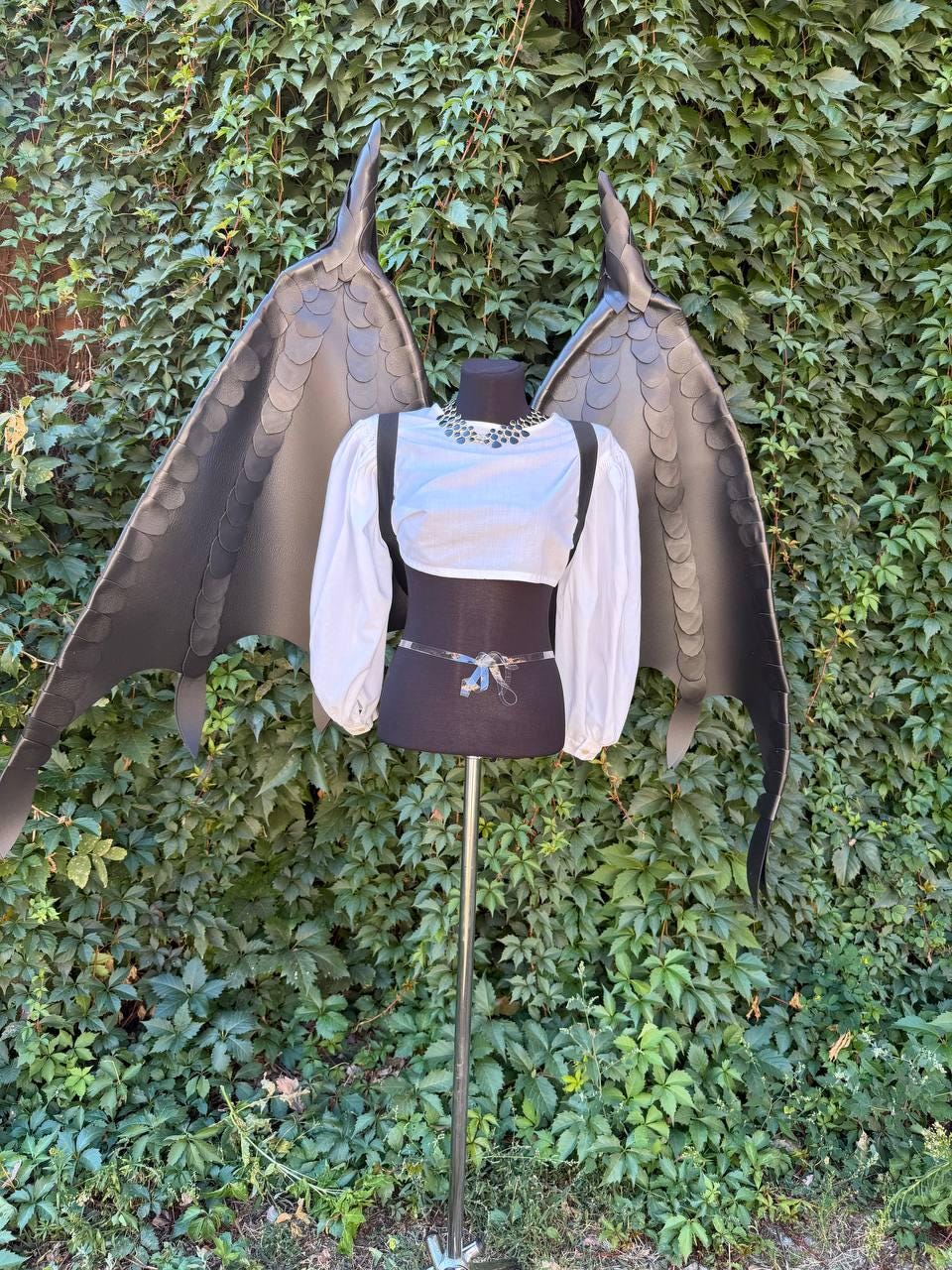 May include: A white long-sleeved cropped top with a high neckline and black harness straps, paired with large, articulated black dragon wings featuring a layered scale texture, displayed on a mannequin torso against a green leafy background.
