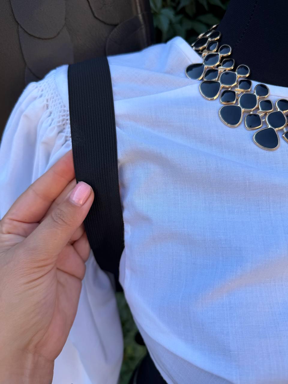 May include: A white blouse with gathered shoulder detailing and a black elastic strap, paired with a statement necklace featuring black, teardrop-shaped enamel pieces set in a gold-toned metal frame that rests against the neckline.