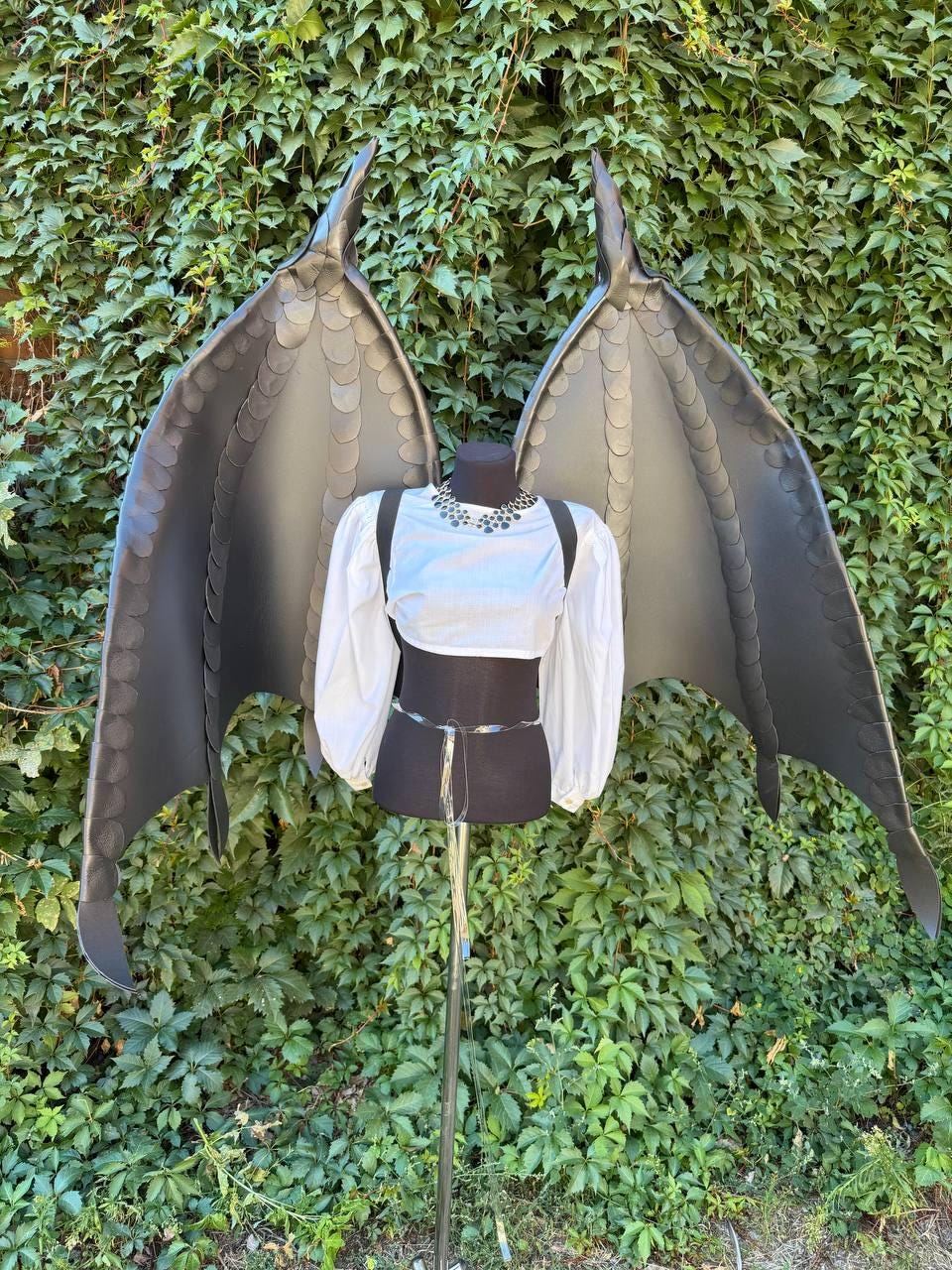 May include: Large, dark gray dragon-style wings with a layered, scale-like texture along the edges, mounted on a black mannequin torso. The mannequin wears a white, long-sleeved cropped top with puffed sleeves and a silver-toned beaded necklace.