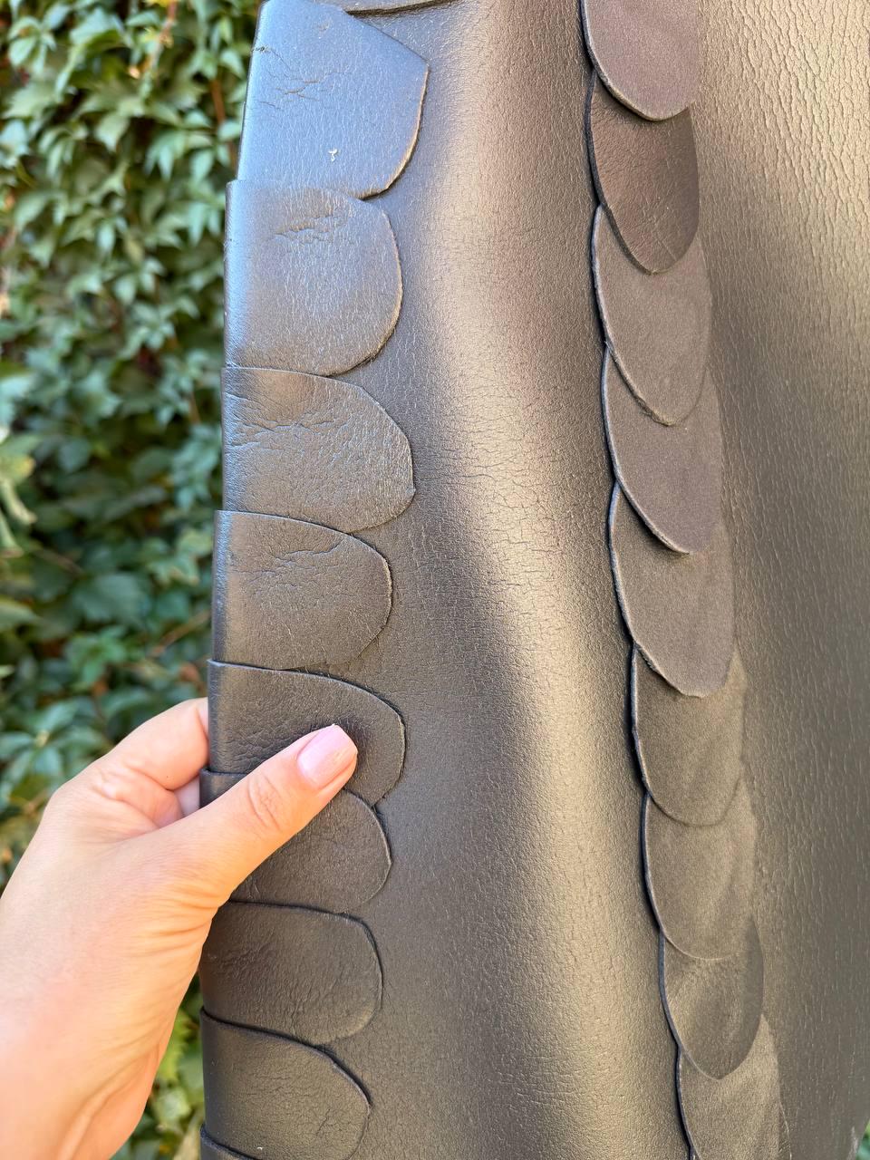 May include: A dark gray or black item featuring a textured surface with two vertical rows of overlapping, rounded, petal-like leather pieces. The material has a visible grain and a matte finish, with the layered segments creating a distinct pattern.