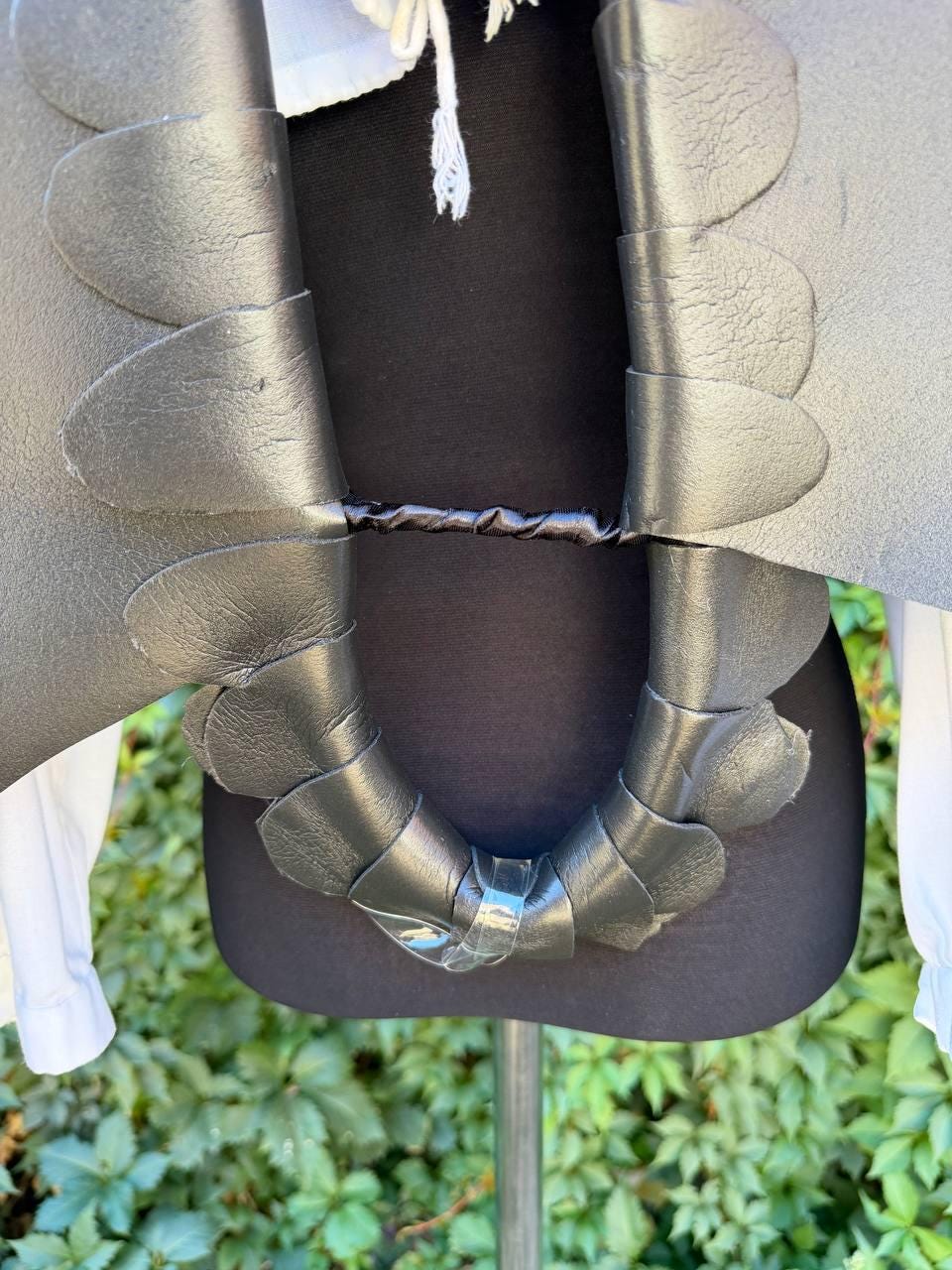 May include: A black leather collar featuring layered, petal-shaped segments that drape around the neck. The collar is secured with a central horizontal strap and a clear plastic fastener at the bottom, worn over a white shirt on a mannequin.