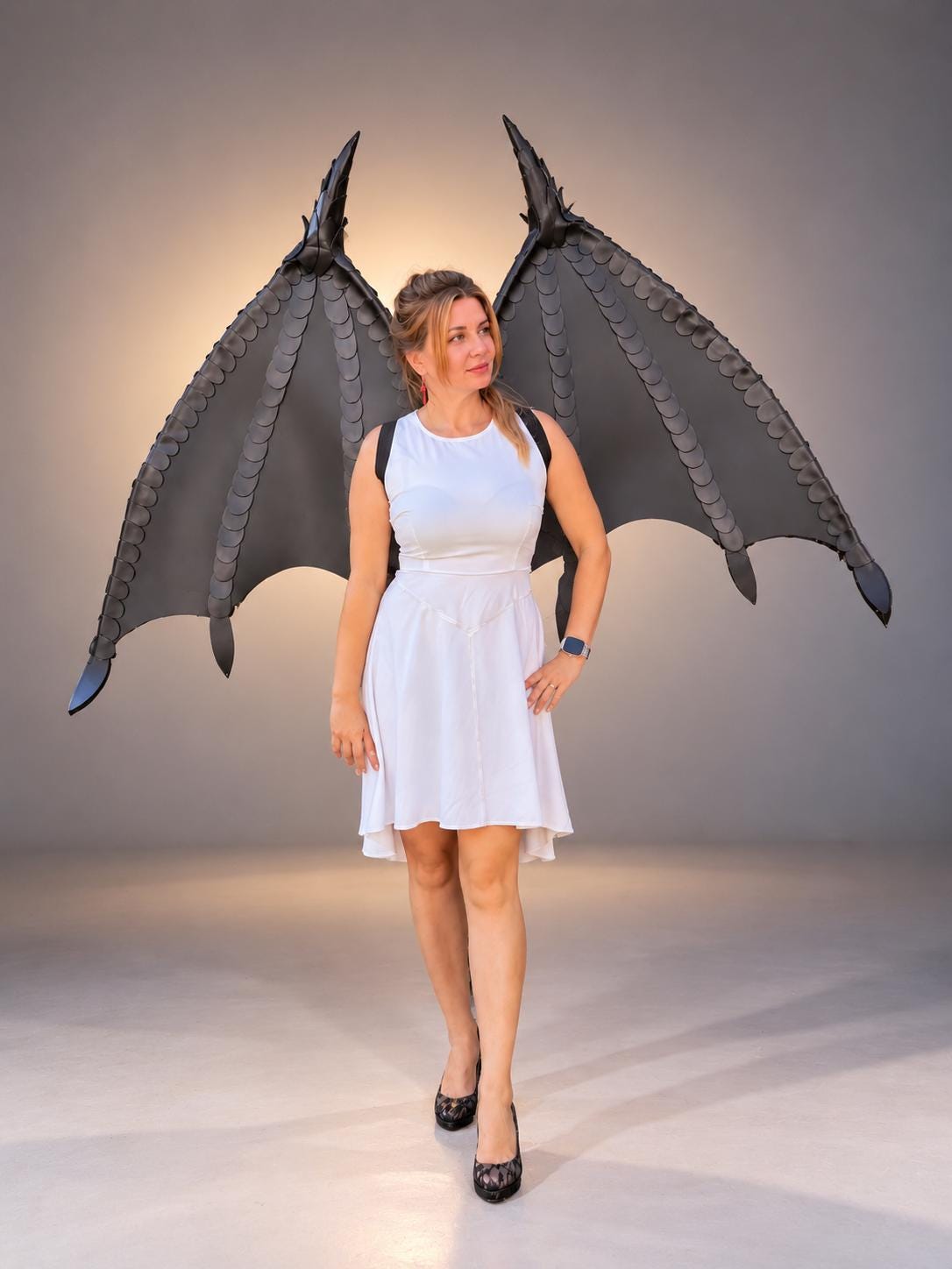 May include: A white sleeveless dress with a high neckline and a flared skirt, paired with large, black, articulated dragon-style wings worn on the back with black straps. The wings feature a layered, scale-like texture and pointed tips.