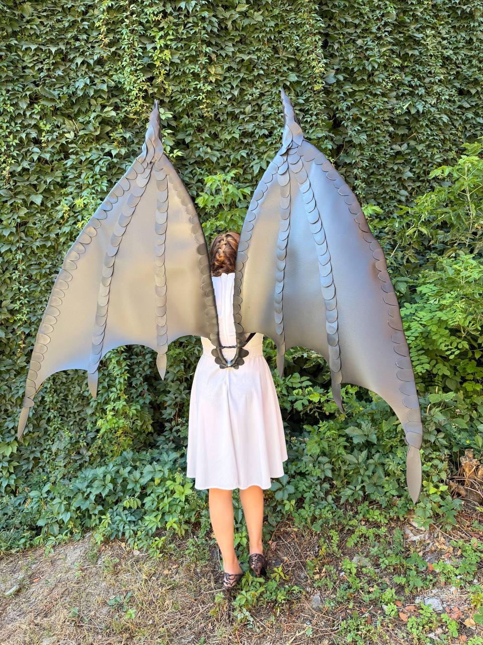 May include: Large, dark gray dragon wings featuring a structured, layered scale design along the edges and spine. The wings are attached to a harness worn over a simple white dress, extending upward and outward with pointed, bat-like tips.