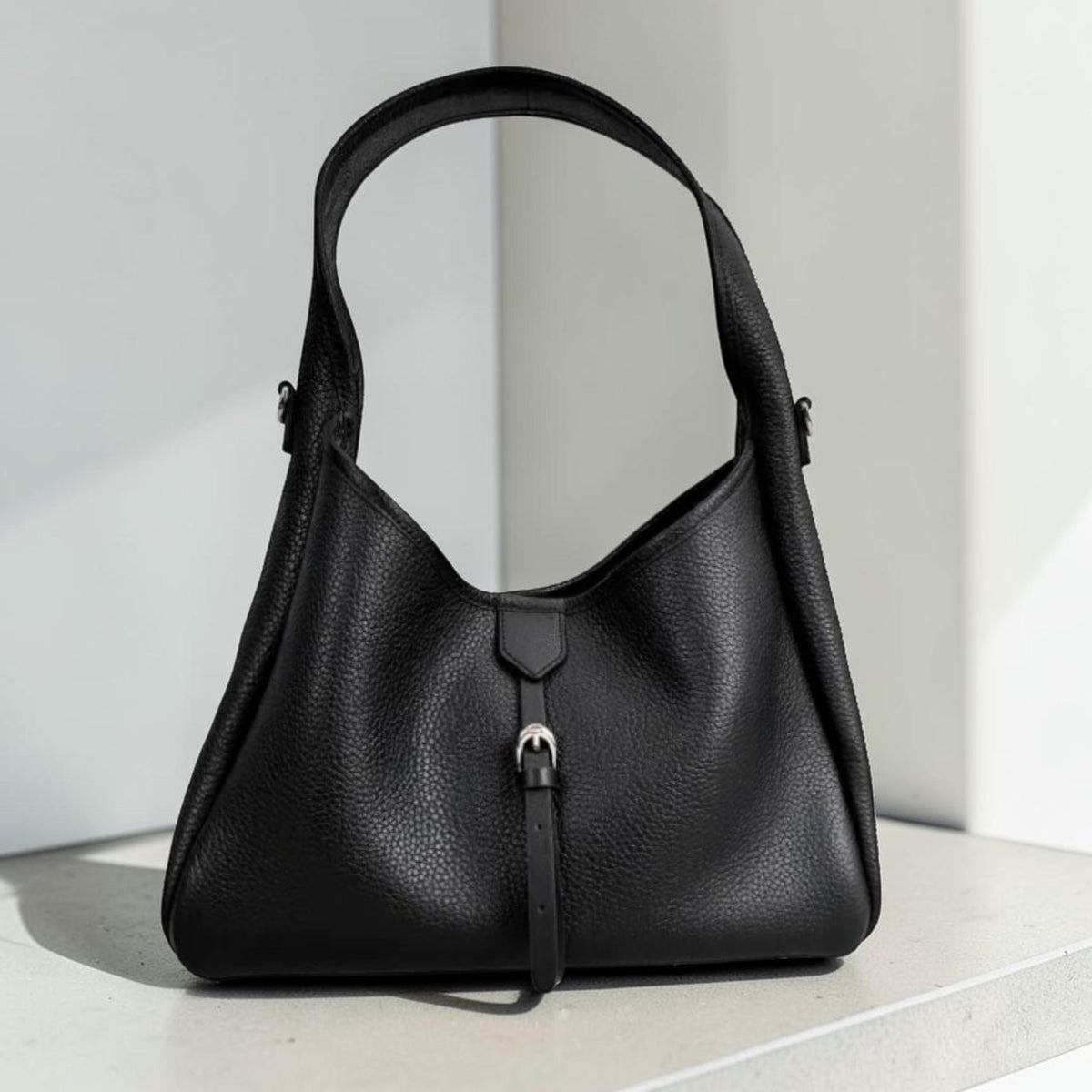 May include: A black leather handbag with a shoulder strap. The bag has a textured surface and a silver-coloured buckle closure. The bag is a trapezoid shape with a curved top.