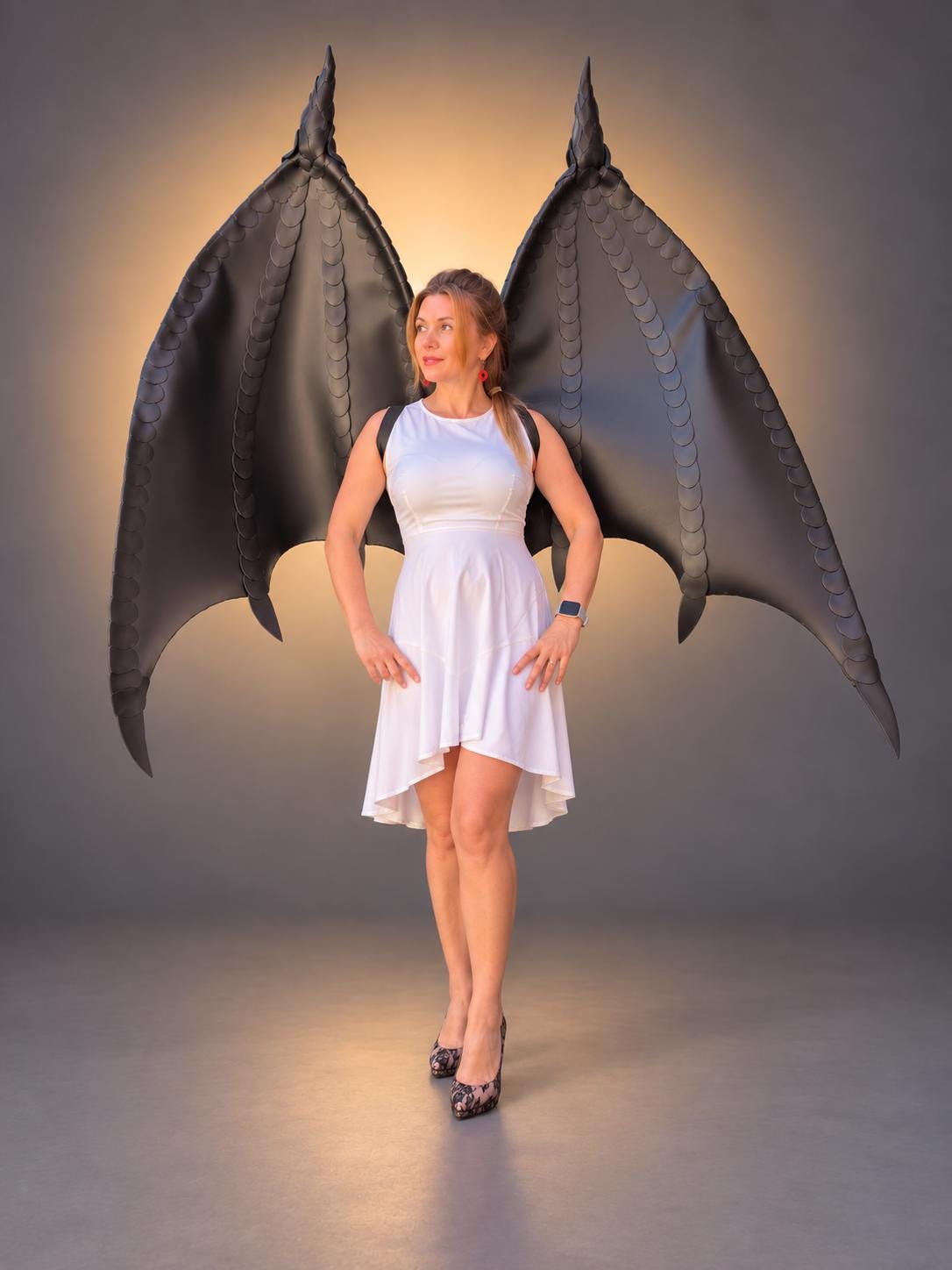 May include: A pair of large, black, articulated dragon wings with a textured, scale-like pattern along the edges. The wings are mounted on a harness structure worn over a white, sleeveless, high-low dress with a fitted bodice and flared skirt.
